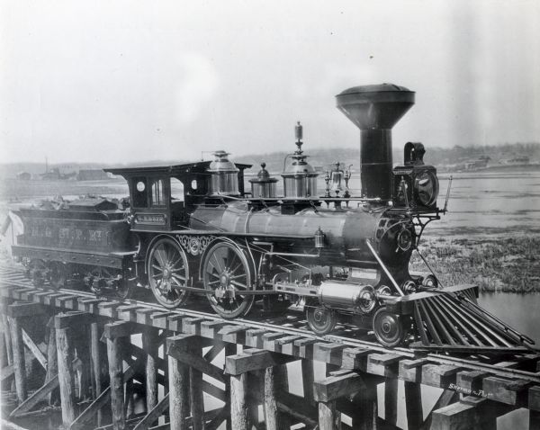 View of one of the first locomotives of the Fox River Valley, Milwaukee and St. Paul Railroad on a wooden trestle over a wetland. The locomotive has brass trim and pulls a wood car.