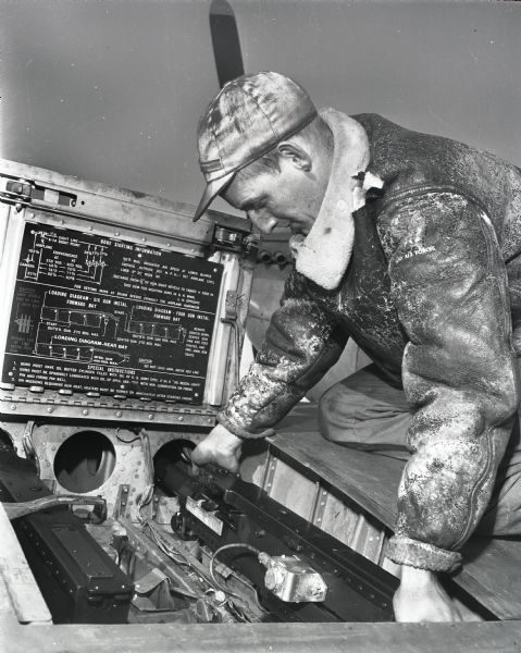 Wisconsin Air National Guard. A man in a leather jacket and cap is loading (?) guns in wings of an F-51 fighter plane.