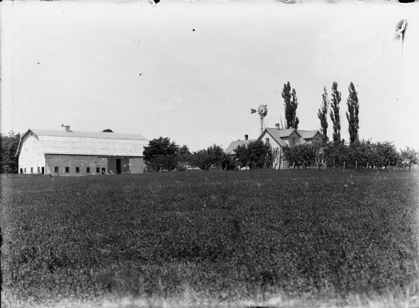 View of a farm taken from a field. Barn, farmhouse, and a windmill are visible behind trees, perhaps an orchard.