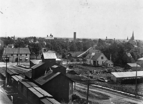 View over a train stopped at a grain elevator in the foreground. The Hotel Ristau is across a street on the left, and a church building is in the distance on the right.