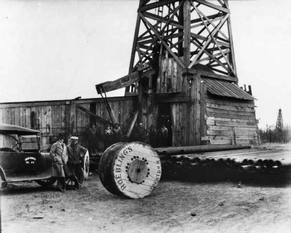 Dr. Herbert Tanner and Ross Moorhead standing in front of an an Okeh Ranger Oil Co. car. There is a large wooden spool in the foreground stamped Roebling's, Trenton, NJ. There are also several pipes piled at right and a small group of workers standing outside an oil well at the Gordon Petroleum Company Ranger Oil Field.
