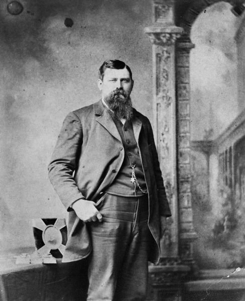 Studio portrait in front of a table and painted backdrop of a bearded George F. Pond. He is wearing a suit coat, vest, and trousers, and has a watch fob chain hanging from his vest.