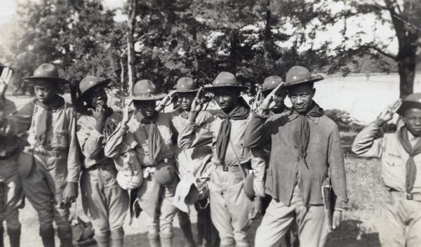 Group of Boy Scouts outdoors in uniform and saluting. There is a lake in the background.
