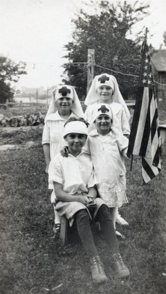 Outdoor portrait of three girls dressed as Red Cross nurses and a boy with a bandage on his head. One girl holds a U.S. flag.