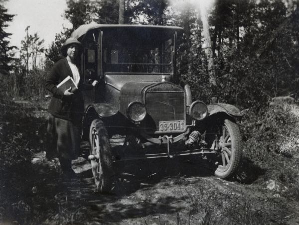 Theta Mead standing next to her car on a rural dirt road holding a book under her arm.