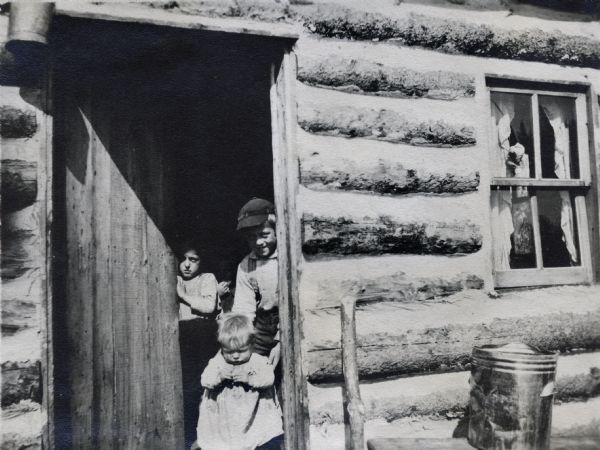 Three children peek out of the door of a log cabin. There is an ash can to the right of the door.