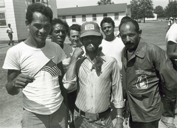 Group of six men posed for an informal portrait. One smokes a cigarette and has a pet squirrel on his shoulder. Another man holds a small U.S. flag. Barracks are visible in the background. Photographs made on July 4, 1980, by Archibald of Cuban refugees who had arrived as a result of the Port of Mariel exodus, and were housed at Camp McCoy, Wis.; including images of the camp life of Cuban men, women, and children.