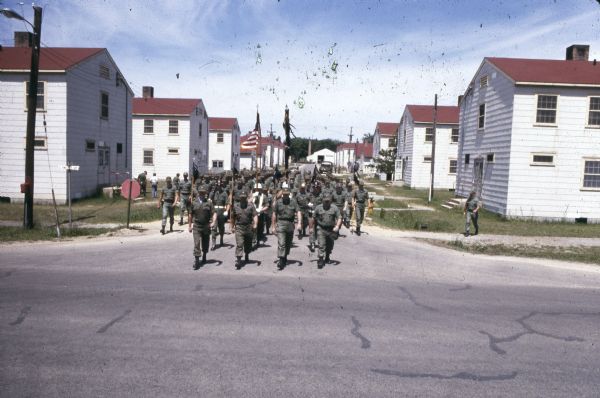 Troops in uniform march on a street at Camp McCoy. There are barracks on either side of the road. One soldier carries a U.S. flag while others carry smaller unidentified flags.