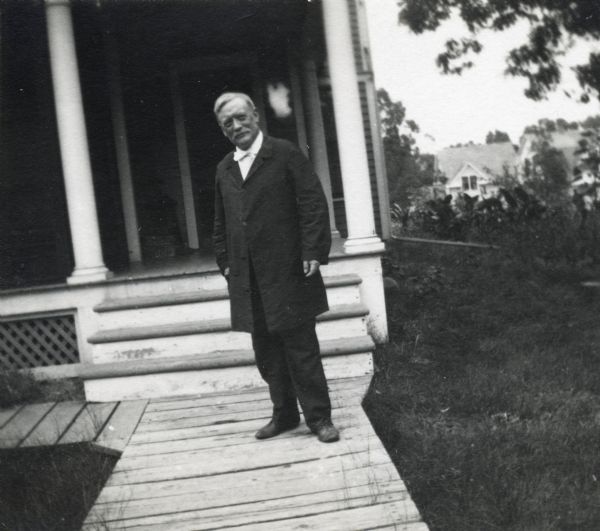Informal full-length portrait of Reverend John Nelson Davidson standing on a wooden sidewalk. He stands at the foot of the stairs of the front porch of a home.