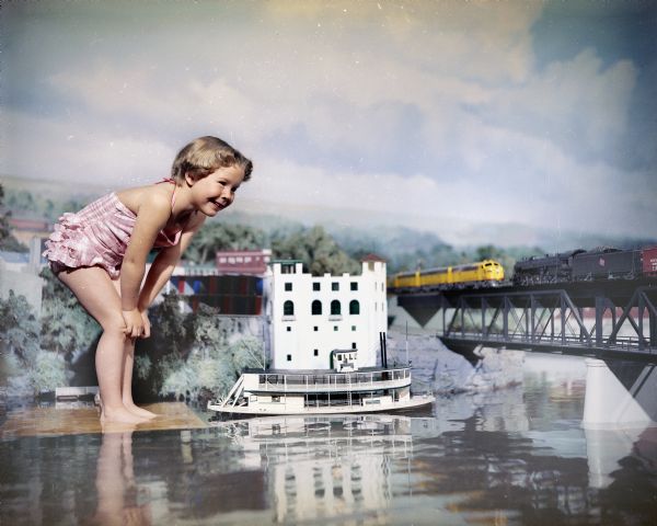 Young girl in a bathing suit standing on a platform in the water looking over the Minirama scale model of Dells Landing. Also in the photograph are a miniature steamboat, powerhouse, and railroad bridge with model trains.