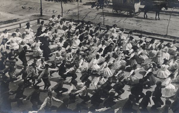 Elevated view of a large group of school children exercising in the schoolyard. Boys and girls are on separate sides of the yard. All are the students are crouching with their arms extended. The teacher is standing at the center toward the back of the group. There is a man standing in a covered horse-drawn wagon on the street in the background.