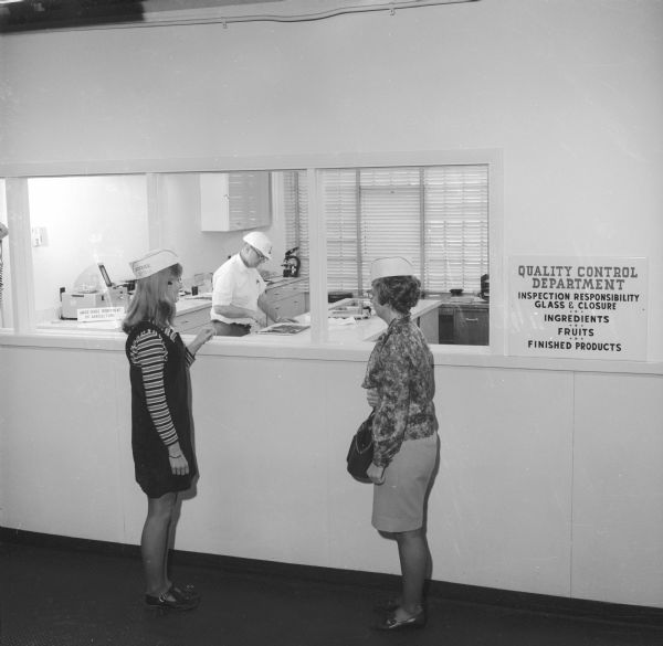 Two women in paper "Smucker's" hats are watching a man at work in a laboratory beyond a set of windows, presumably at J.M. Smucker Company's Quality Control Department. The man, wearing a hard hat, appears to be examining a jam-like substance on a tray. A sign bearing the words "United States Department of Agriculture" sits before some kind of computerized equipment. A sign besides the windows states, "Quality Control Department Inspection Responsibility Glass & Closure; Ingredients; Fruits; Finished Products."