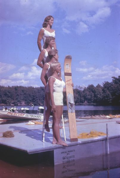 Four female water skiers posed in profile in white bathing suits on a pier or floating raft. Three stand on a ladder so they appear in a vertical column. One of the women holds up a water ski. There is a man in a boat in the background with Mercury outboard motors.