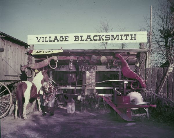 A man in a plaid shirt and heavy apron holding the hoof of a horse in one hand, and a long file in the other. They are surrounded by metal wares and metal working tools in an open shed beneath a large sign reading "Village Blacksmith." There is a plow at right.