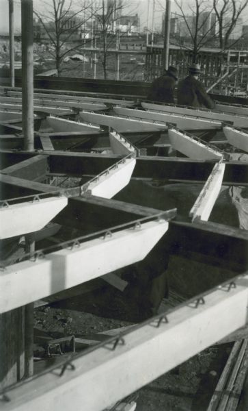 A construction detail in the House of Tomorrow showing the red top floor joists in place. Two men wearing hats are on the structure with their backs to the camera looking down. In the background are other structures and buildings.