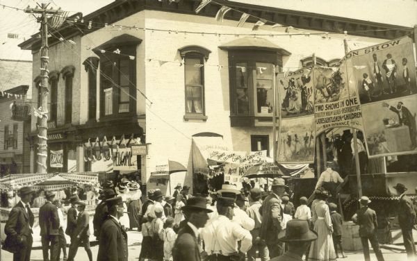 Crowd near the front of a side show tent with large banners.