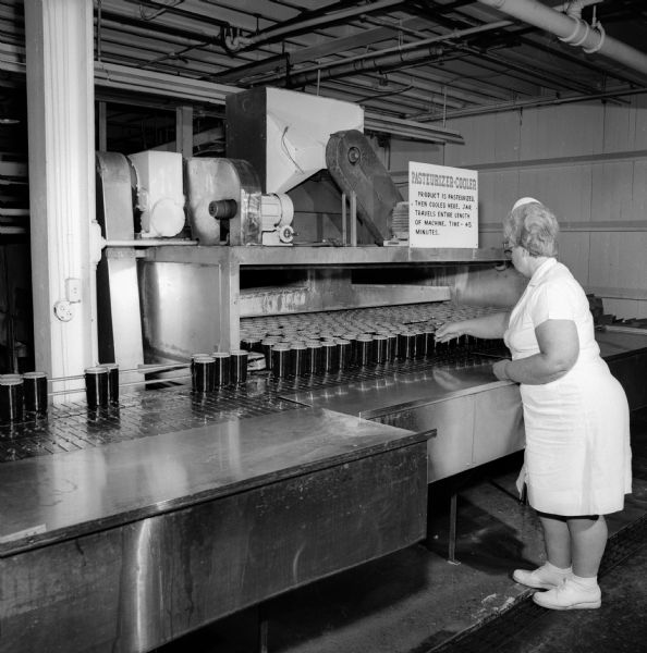 A woman wearing a hairnet and a white smock reaches towards jars of jam or jelly that appear to be coming off a piece of factory equipment labeled "Pasteurizer-Cooler" at a J.M. Smucker Company plant.  The sign states that the "product is pasteurized, then cooled here. Jar travels entire length of machine. Time - 45 minutes."