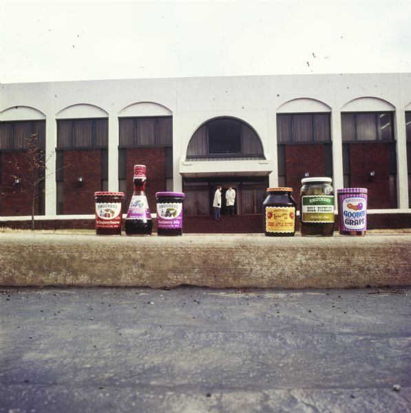 A display of some of the J.M. Smucker Company products. The products are displayed in front of the company's corporate headquarters. In the background, two men are standing on steps in front of the entrance.