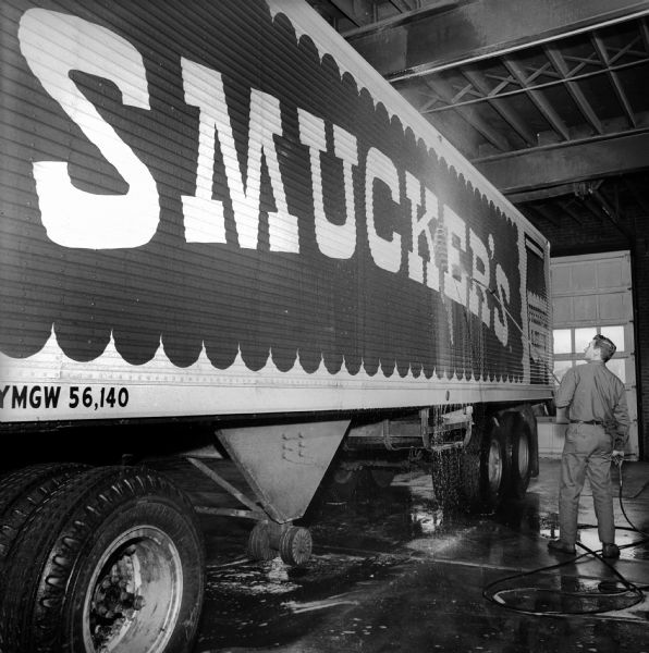A uniformed man in an indoor garage or warehouse is using a power washer to clean a truck trailer emblazoned with the Smucker's logo.