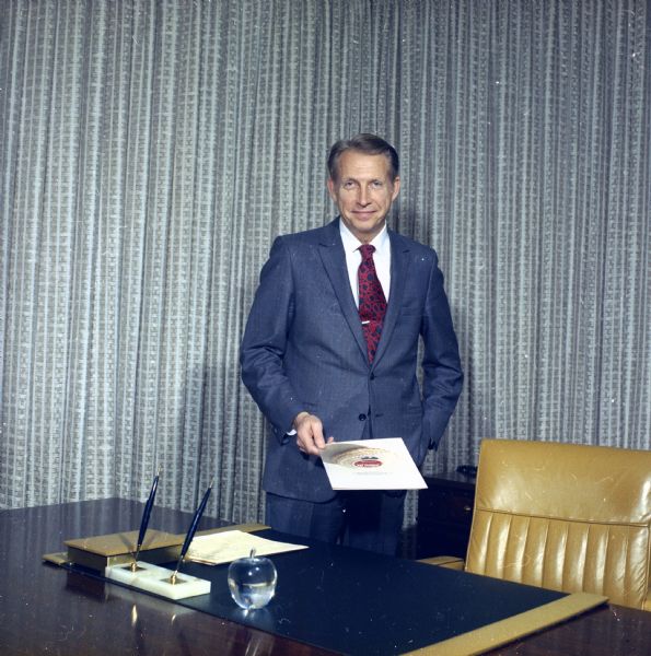 Portrait of J.M. Smucker Company executive Paul Smucker standing next to his desk holding a brochure.