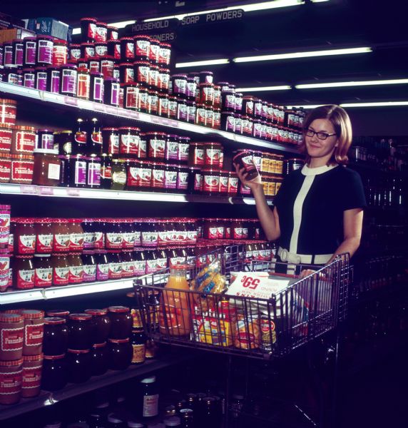 A publicity photograph of a bespectacled young woman, in a 1960s mod-style dress, examining a jar of Smucker's strawberry preserves in an aisle of a grocery store. Visible in the shopping cart in front of her are a box of Lipton tea, a can of green beans, other food items, and some sort of coupon. Visible on the grocery shelf are other Smucker's products as well as "McHenry's Sorghum" on the bottom shelf.