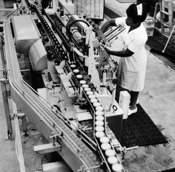Publicity photograph of a female employee, in a white, nurse-like uniform, white nursing cap labeled "Smucker's" and white nursing shoes, operating a labeling machine at a J.M. Smucker's Company plant. The original caption reads: "Parade of filled jelly jars passes through the labeling machine at one of Smucker's three preserving plants. Alternate track is a bypass around labeler for use in case of machine problems."