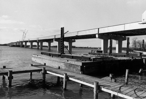 View from the east shoreline of construction of the bridge over Little Lake Buttes des Morts. There is a pier and what appears to be a barge in the foreground. Two cranes are on another barge in the middle of the semi-completed bridge.