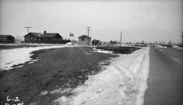 View of a gravel road next to U.S. Highway 41 with buildings in the background. There is a sign for "Fox Tavern," and a car is parked in front of a small building. The log building on the left has a sign for "Standard Service."