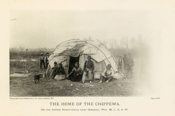 Chippewa Indians are seated around a dome-shaped dwelling on a reservation.