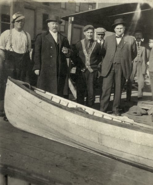 Billy Hughes in a long coat and hat and smoking a cigar stands with five other men in front of a boat at Schott's Landing.