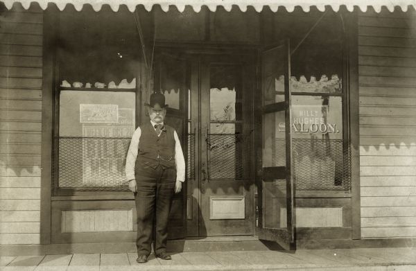 Billy Hughes in a vest and bowler hat standing in front of his saloon.