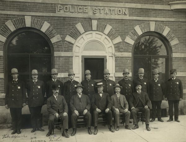 Group portrait of police officers standing and others seated in front of the Police Station.