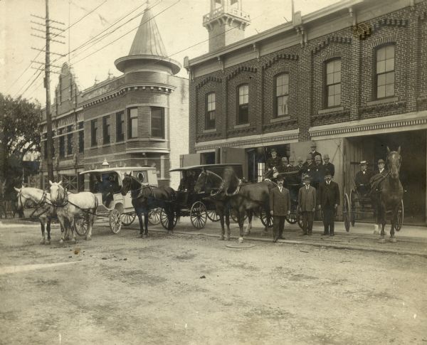Police and others shown posed in and around horse-drawn carriages in front of a large brick building with open doors. The City Ambulance is on the left drawn by two horses. The building on the left in the background is the Police Station.