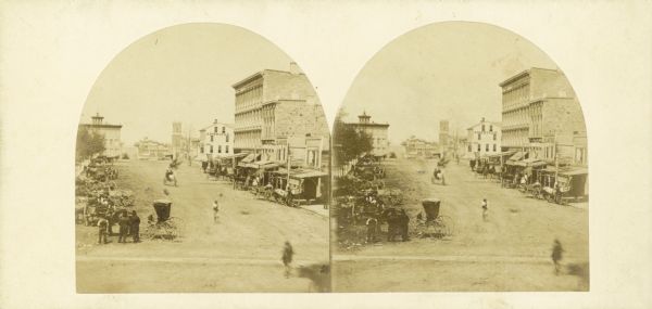 Stereograph of Pinckney Street, about 1860, from the roof of the Fairchild Block at the corner of East Main and South Pinckney looking northwest toward Lake Mendota. Bruen's Block is the large commercial building on the right. Next to it, across the East Washington intersection, is the historic American House hotel where the Legislature first met. On the left numerous horse-drawn buggies and wagons are hitched along Capitol Park fence. Bacon's Block is the building with the turret on its roof at the corner of Pinckney and Mifflin streets.