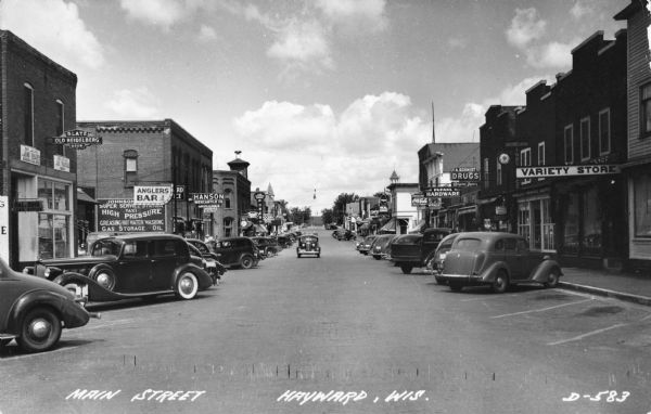 A view down Main Street with cars parked at an angle in front of storefronts. Caption reads: "Main Street, Hayward, Wis."