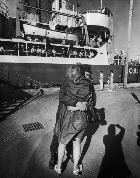 A returning soldier hugs a woman on the dock, who is probably his girlfriend or wife. A military (naval?) ship is visible in the background.