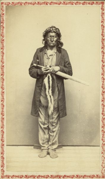 Carte-de-visite studio portrait of a Ho Chunk chief standing in front of a backdrop. He is wearing a long coat, fringed shirt, and a crown on his head. In his hands he carries a long-stemmed pipe and a skunk tobacco bag.