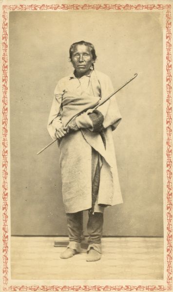 Carte-de-visite full-length studio portrait in front of a backdrop of the Winnebago (Ho-Chunk) chief. In this portrait he is past the age of seventy. He is standing erect, though he is carrying a carved cane in his arms. A photographer's stand is directly behind him.