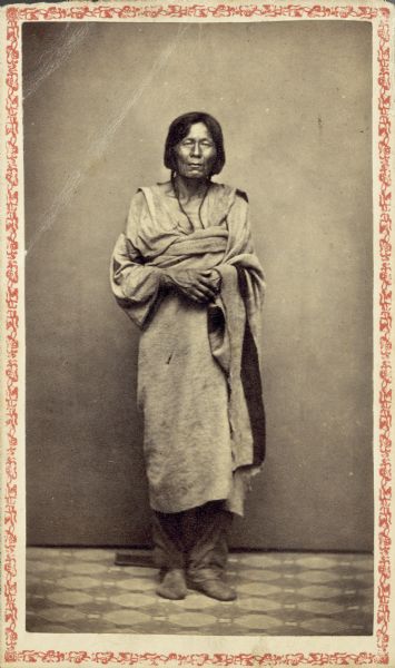 Carte-de-visite studio portrait of Winnebago (Ho-Chunk) man standing in front of a backdrop. He is wearing a blanket over a shirt and trousers.