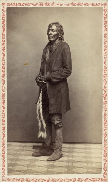 Carte-de-visite studio portrait of Winnebago (Ho-Chunk) man standing in front of a backdrop. He is wearing a long coat, trousers, and boots, and is holding skunk skin tobacco bag.