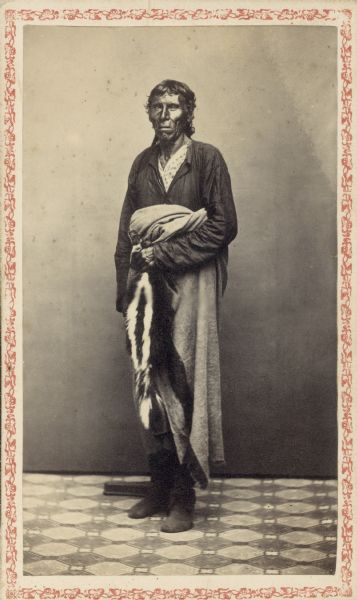 Carte-de-visite studio portrait of Winnebago (Ho-Chunk) chief standing in front of a backdrop. He is wearing a coat, shirt, and trousers and is holding a blanket and a tobacco bag made from the cased skin of a skunk.
