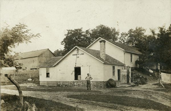 Exterior of Brodhead Cheese Factory with man standing in front. The first Jaggi family factory was known as County Line Co. The business shipped cheese in barrels, which were at first stored in the open.