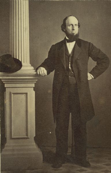 Full-length studio portrait of Daniel Wells, who is standing next to a column with his fist on his hip. His hat is resting on the column.