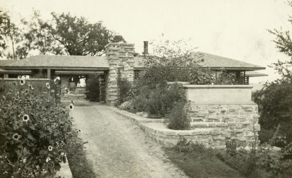 The entrance to Taliesin showing a driveway with a roofed entrance. Flowers and plants border the drive. Taliesin is located in the vicinity of Spring Green, Wisconsin.