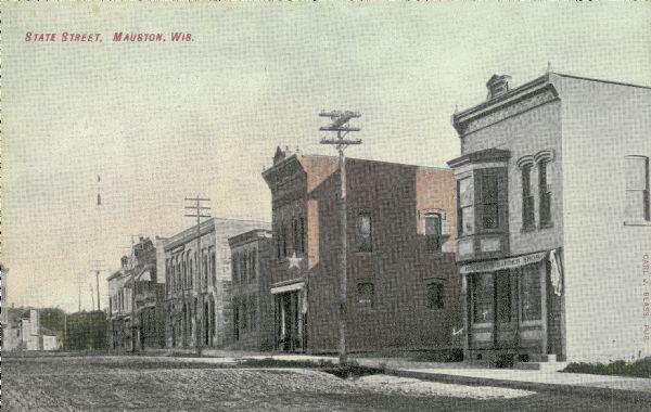 View across State Street showing storefronts along the right. Star sign shown on building was donated to museum. Caption reads: "State Street, Mauston, Wis."