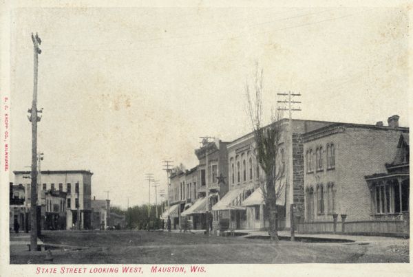 View down wide street with storefronts on both sides. Caption reads: "State Street looking West, Mauston, Wis." Star sign shown on building was donated to museum.