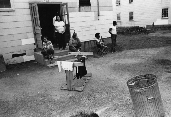 Members of family seated and standing near the entrance to their home. Photographs made on July 4, 1980, by Archibald of Cuban refugees who had arrived as a result of the Port of Mariel exodus, and were housed at Camp McCoy, Wisconsin.