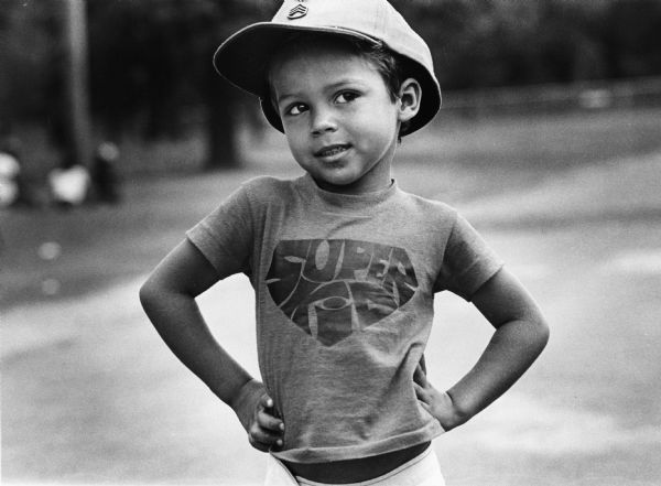 Child posing while wearing a  "Super Kid" tee shirt and a baseball cap. Photograph is part of a group of images taken of Cuban refugees who had arrived as a result of the Port of Mariel exodus, and were housed at Camp McCoy, Wisconsin.