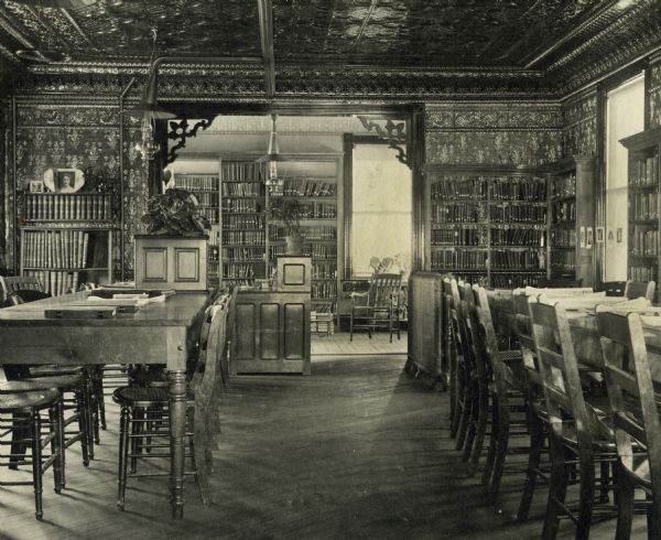 Interior view of the Joseph Mann Public Library, showing the south half of the reading room. The building opened in 1914. Land was funded by a $1000 donation from Mrs. Jos. Mann and a $12,500 donation from Andrew Carnegie. The walls and ceiling are covered with copper plated metal that was a gift from former Two Rivers resident, Hermann Voshardt, of the Freidly-Voshardt Co. of Chicago, manufacturers of metal ceilings. Also in the room are reading tables and chairs, a desk, and bookshelves.