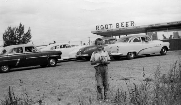 Young boy with a rootbeer float moustache standing in the parking lot of a rootbeer stand.